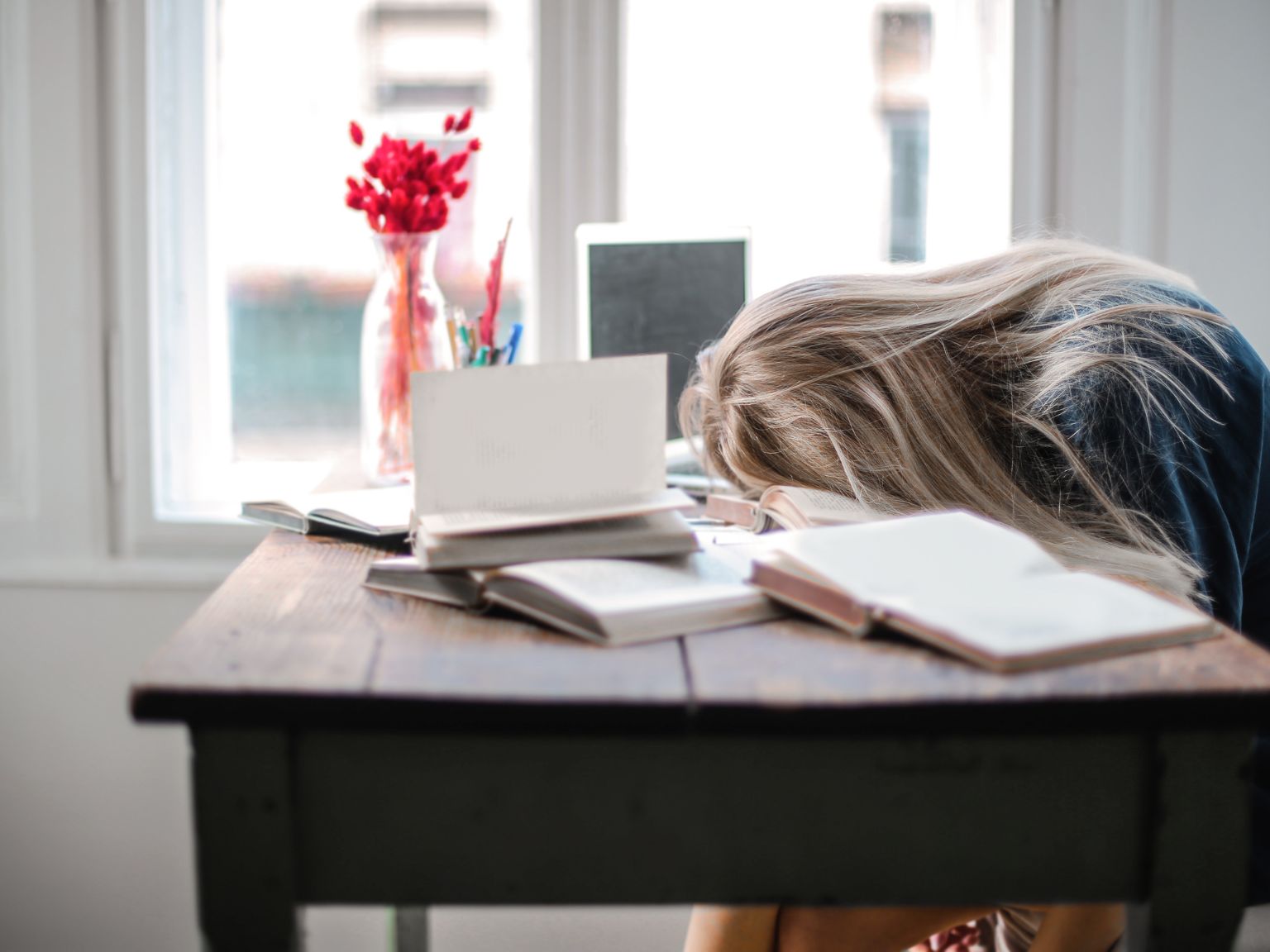 A lady with long blonde hair with her face down on her desk. She is surrounded by open books and her laptop.